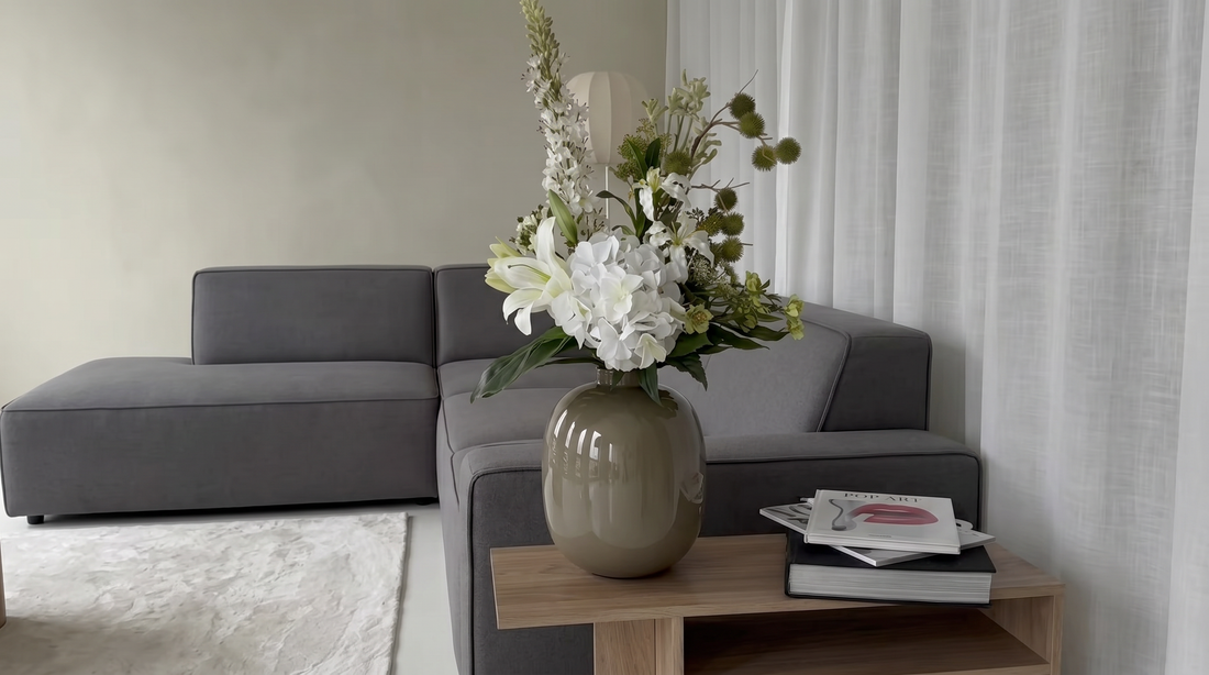 Rustige, moderne woonkamer met een grijze hoekbank, houten salontafel met een vaas vol witte bloemen en groene takken, en enkele tijdschriften naast een groot raam met lichte gordijnen.